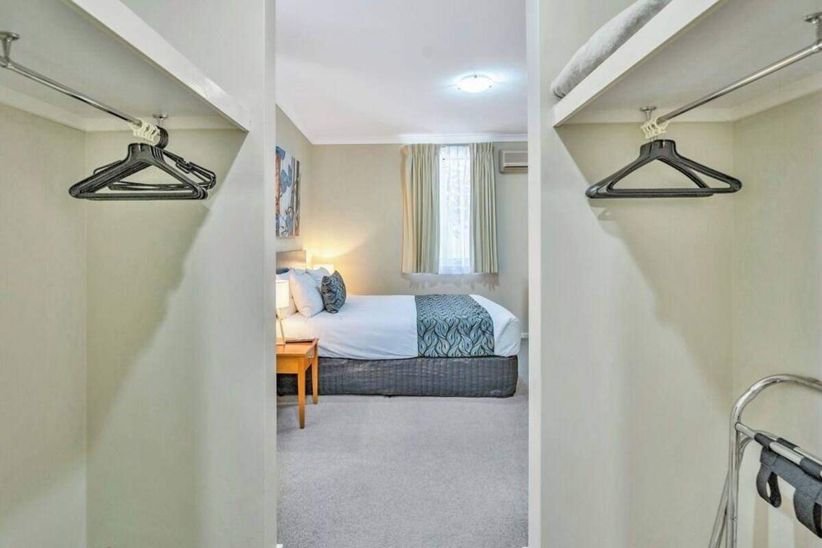 Um corredor estreito que dá para um quarto com cama, mesa de cabeceira e uma janela com cortinas.