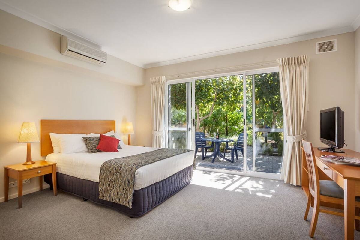 Quarto de hotel com cama, mesa de trabalho, televisão e varanda com área de estar ao ar livre.