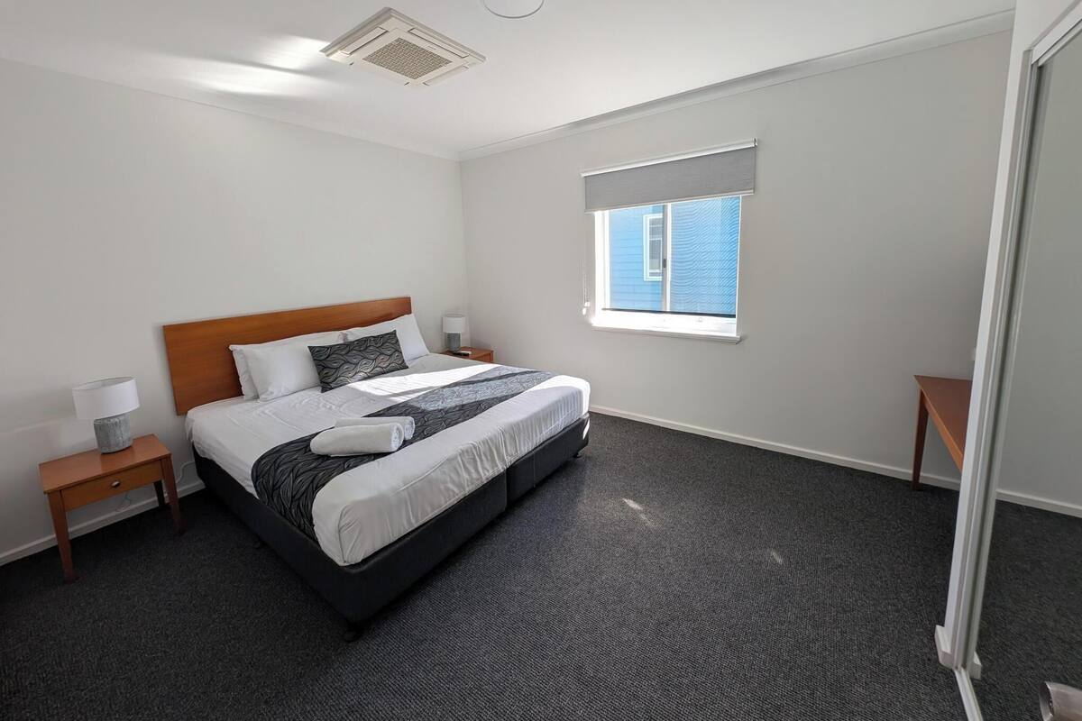 Quarto de hotel com cama, mesa de cabeceira, luminária, janela com persianas e um respiradouro no teto.
