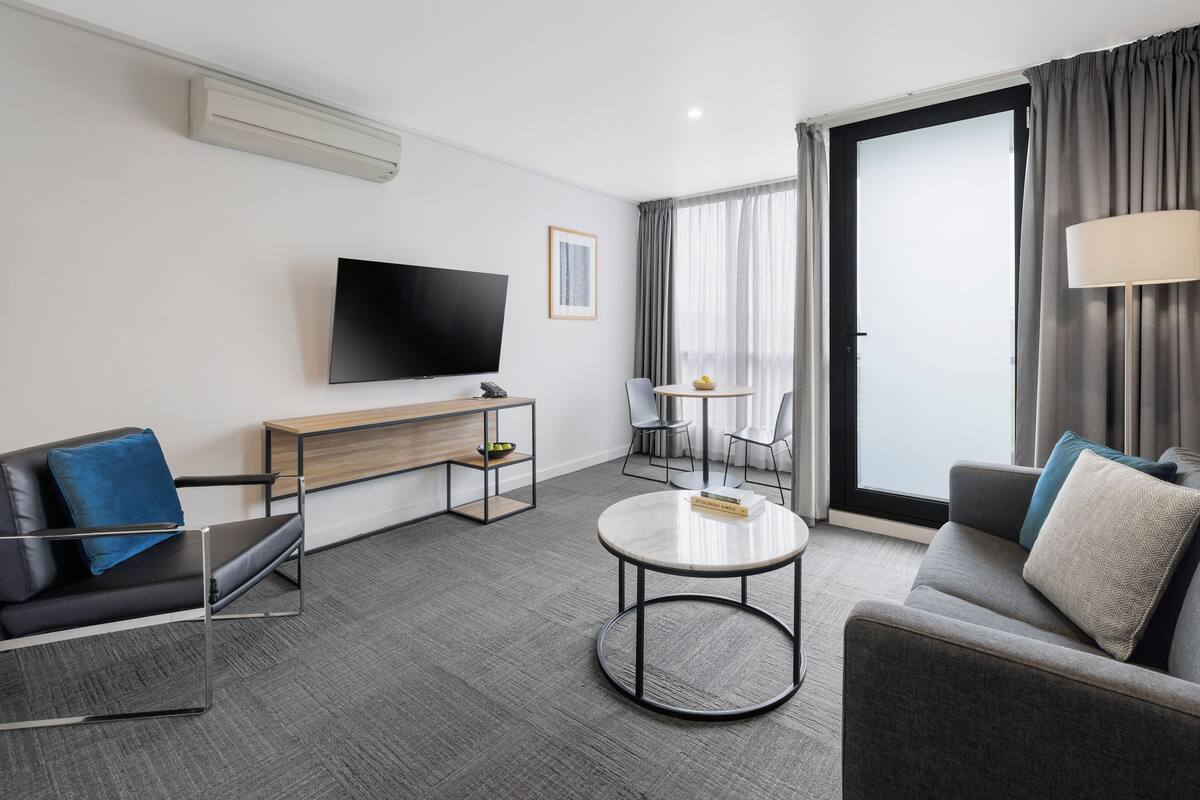A modern hotel room with a flat-screen TV, a sofa, a round coffee table, a desk with a lamp, and a large window with curtains.