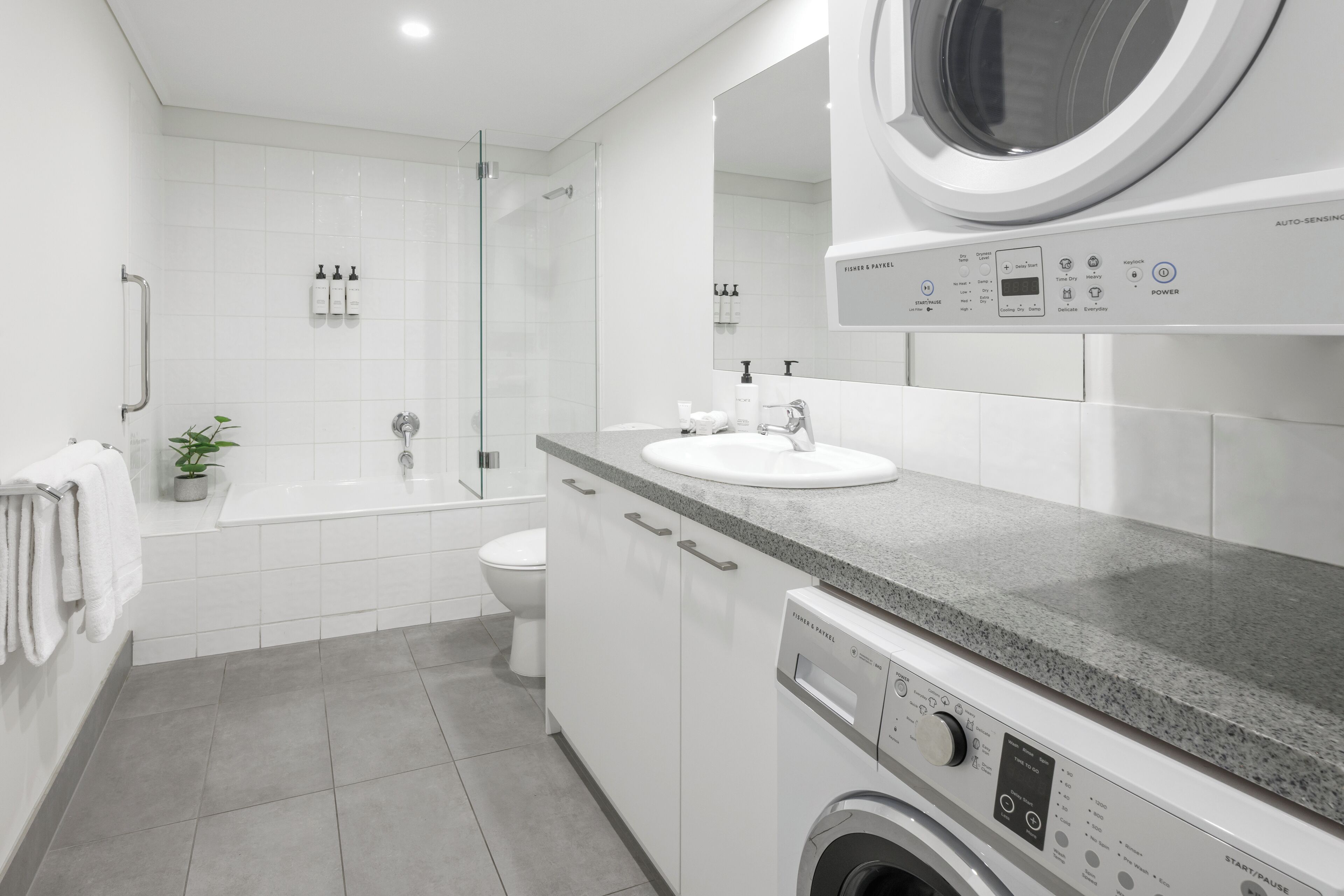 A modern bathroom with a built-in washing machine, a glass shower enclosure, a toilet, and a sink with a countertop.