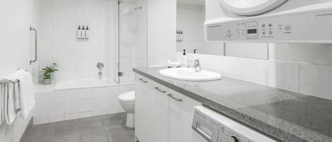 A modern bathroom with a built-in washing machine, a glass shower enclosure, a toilet, and a sink with a countertop.