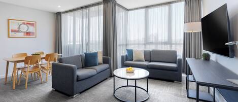 A modern hotel room with a sofa, armchair, dining table, and a flat-screen TV.