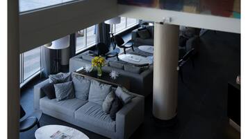 Un área de descanso moderna con un sofá gris, una mesa de centro con un florero y una revista. Se observan varias sillas y una mesa redonda al fondo.