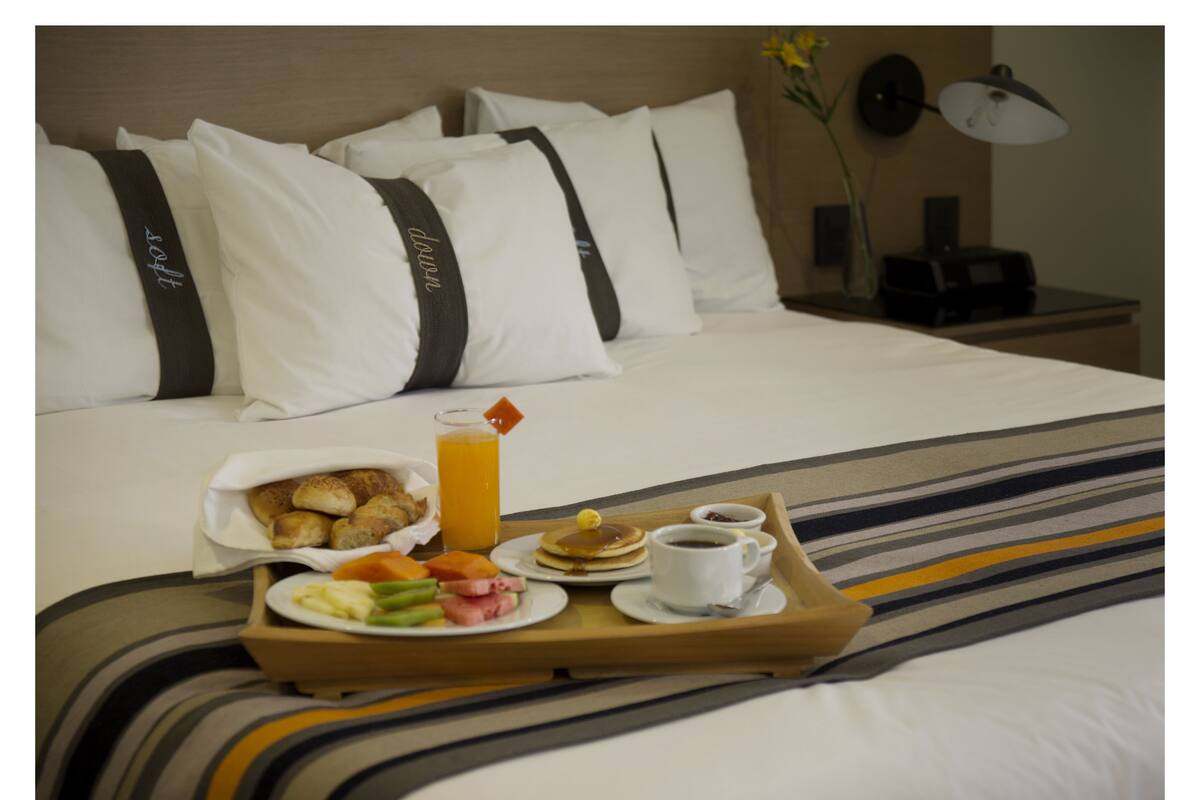A neatly made bed with white and dark pillows, a tray with breakfast including pancakes, eggs, and fruit, and a glass of orange juice.