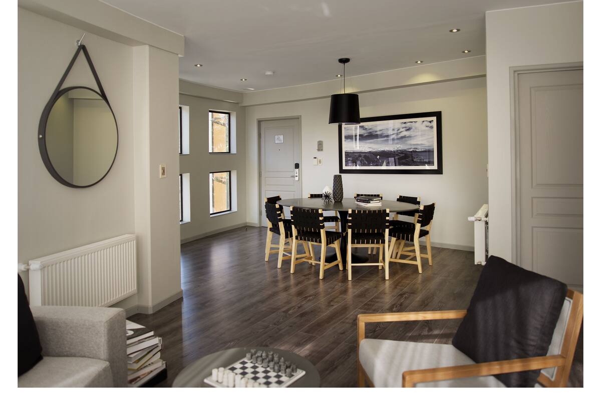A modern living room with a dining area, featuring a round table, chairs, a large mirror, and a cityscape painting.