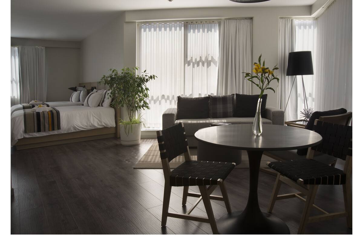 A modern hotel room with a bed, a sofa, a dining table with chairs, a potted plant, and a floor lamp.