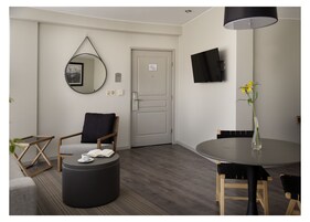 Una habitación moderna y bien iluminada, con una mesa de comedor redonda, una silla cómoda, un pequeño reposapiés, un espejo circular y un televisor de pantalla plana montado en la pared.