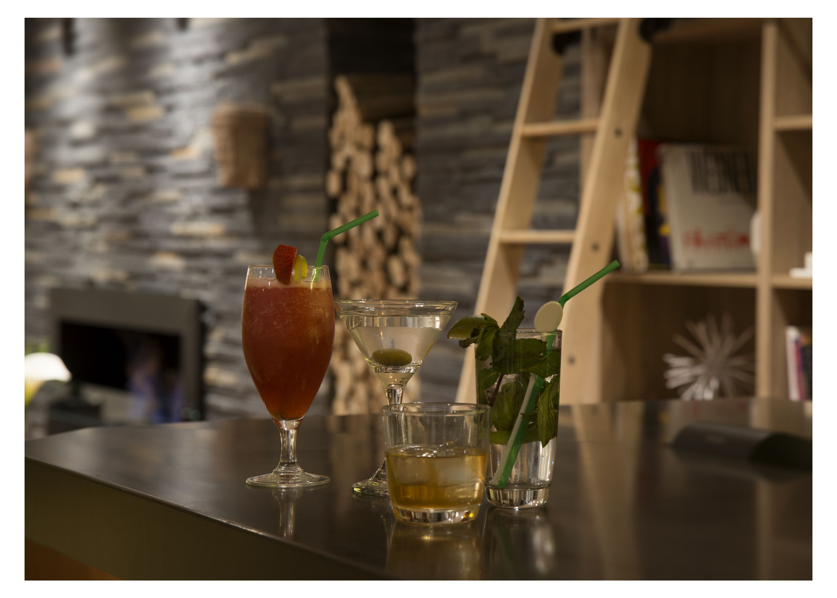 A bar area with three drinks on a counter, a fireplace, and a bookshelf in the background.