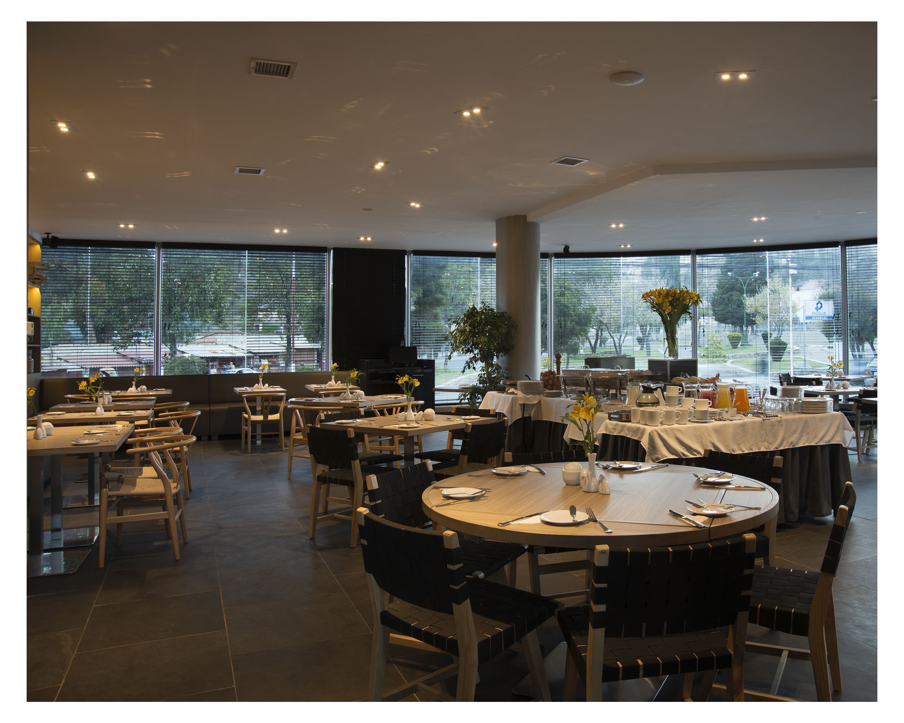 A spacious dining area with multiple tables and chairs, large windows with blinds, and a variety of table settings.