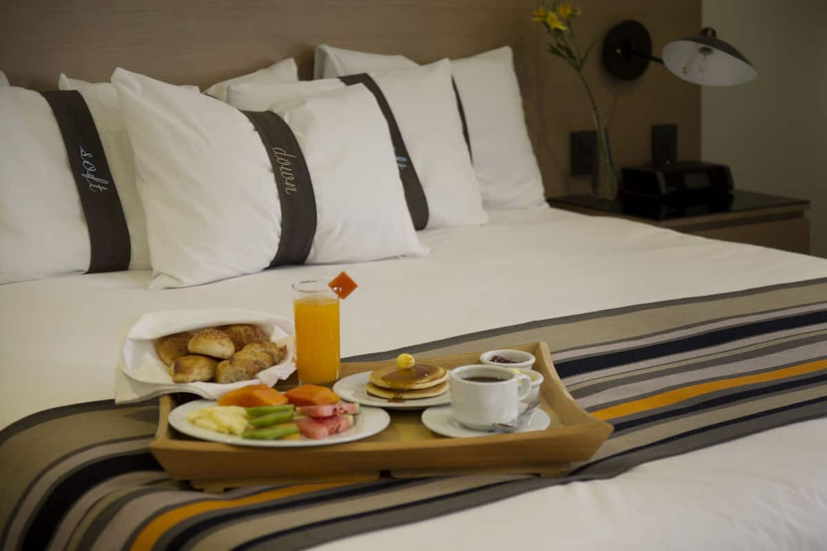 A neatly made bed with white bedding and a striped blanket, a tray with breakfast including toast, fruit, pancakes, and coffee, and a bedside lamp on a nightstand.