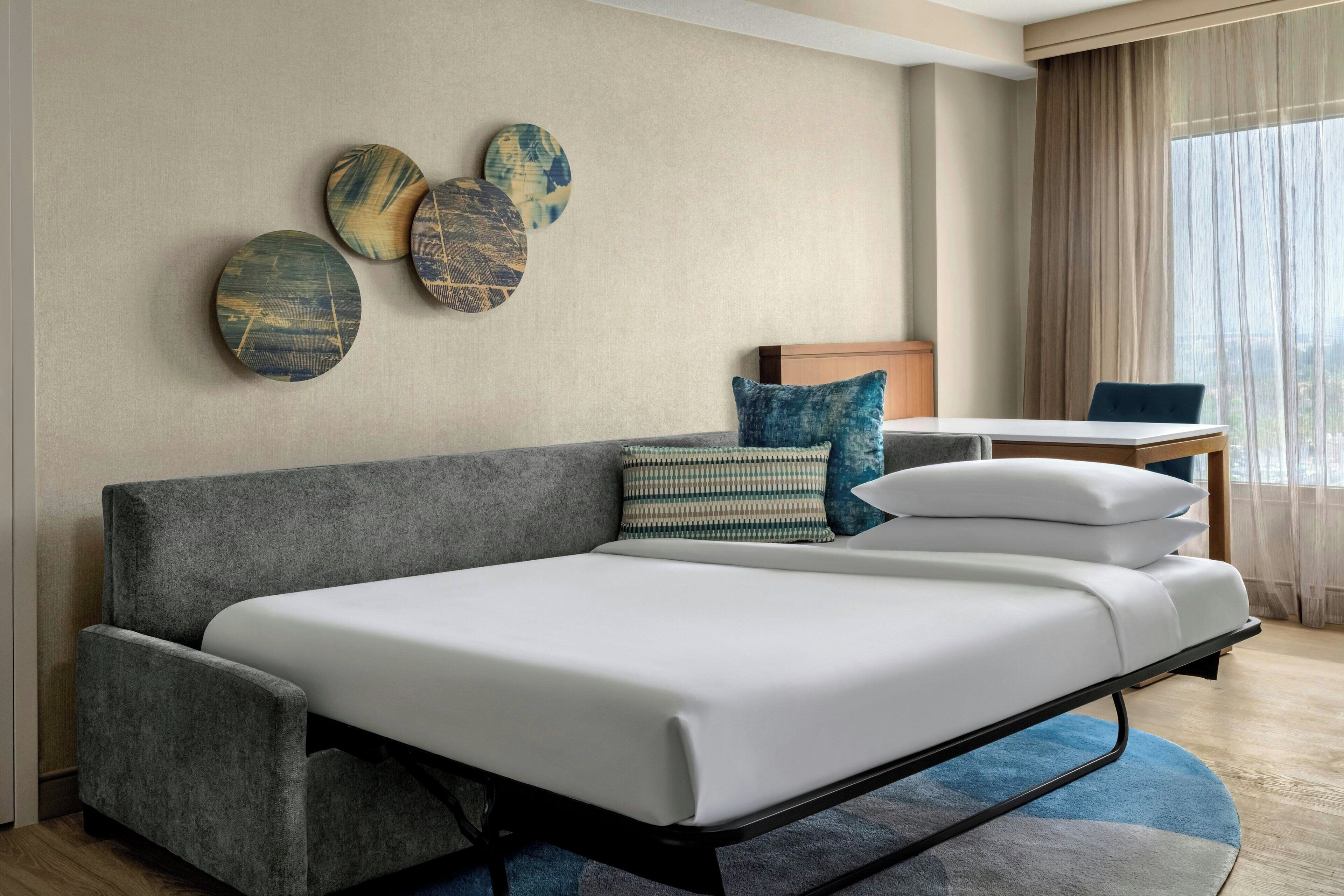 A modern hotel room with a grey sofa, a small table, and a view of the city through large windows.