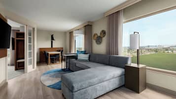 Uma sala de estar moderna com um sofá seccional cinzento, uma área de jantar com mesa e cadeiras, uma TV de tela plana e uma janela grande com vista para a cidade.