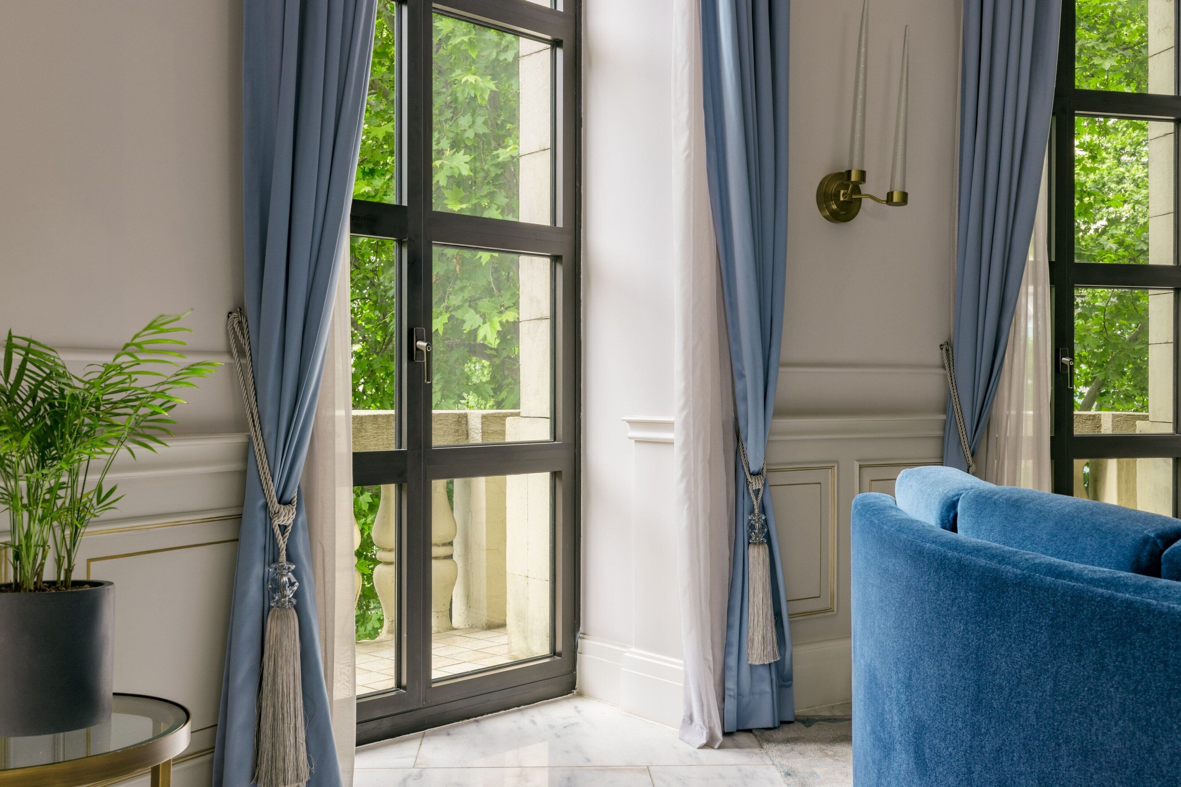Une pièce avec un canapé bleu, une plante en pot et une vue sur la verdure grâce à de grandes fenêtres ornées de rideaux bleus.
