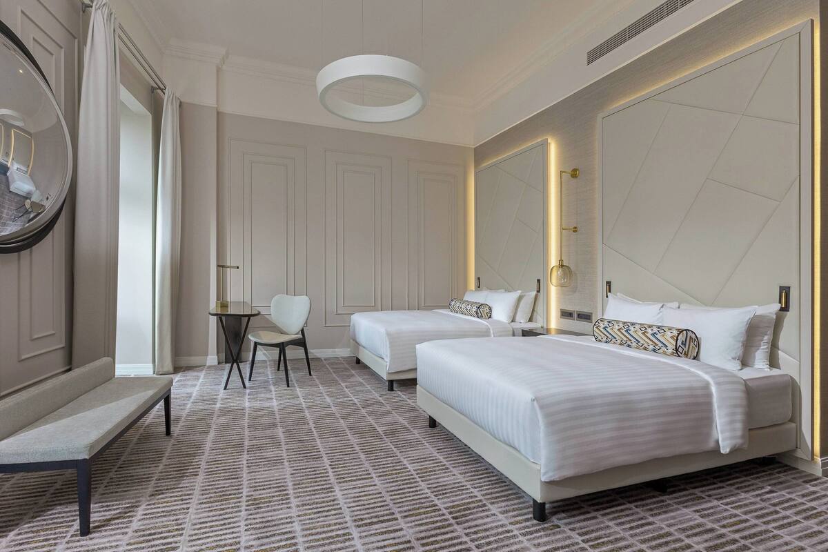 A hotel room with two beds, a bench, a small table, and a mirror.