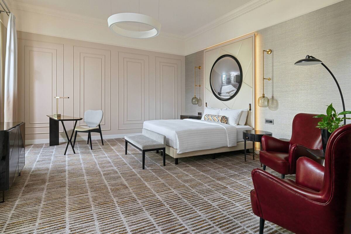A bedroom with a bed, a red armchair, a desk, a chair, a mirror, and a hanging light fixture.