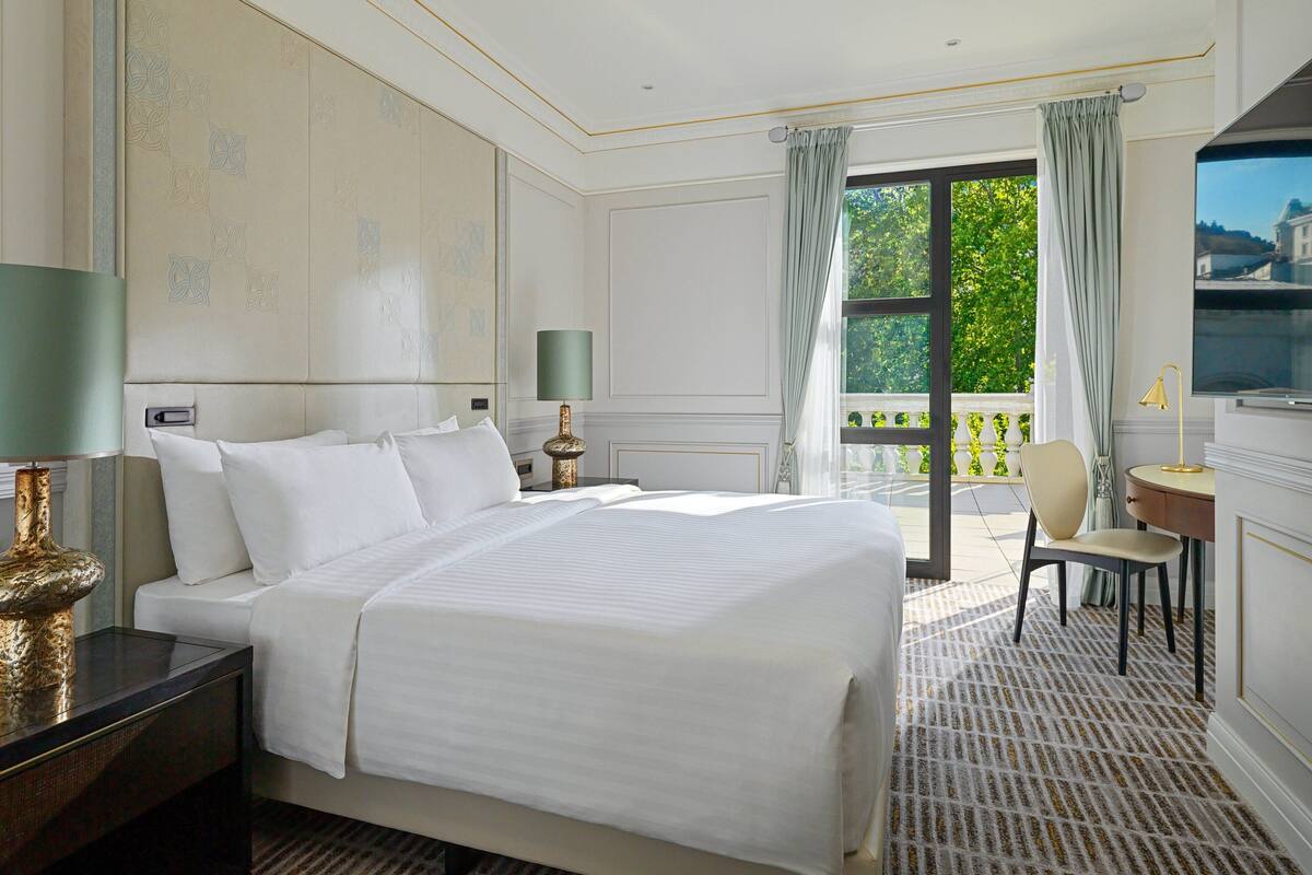 A hotel room with a large bed, a desk with a chair, a TV, and a balcony with a view of greenery.