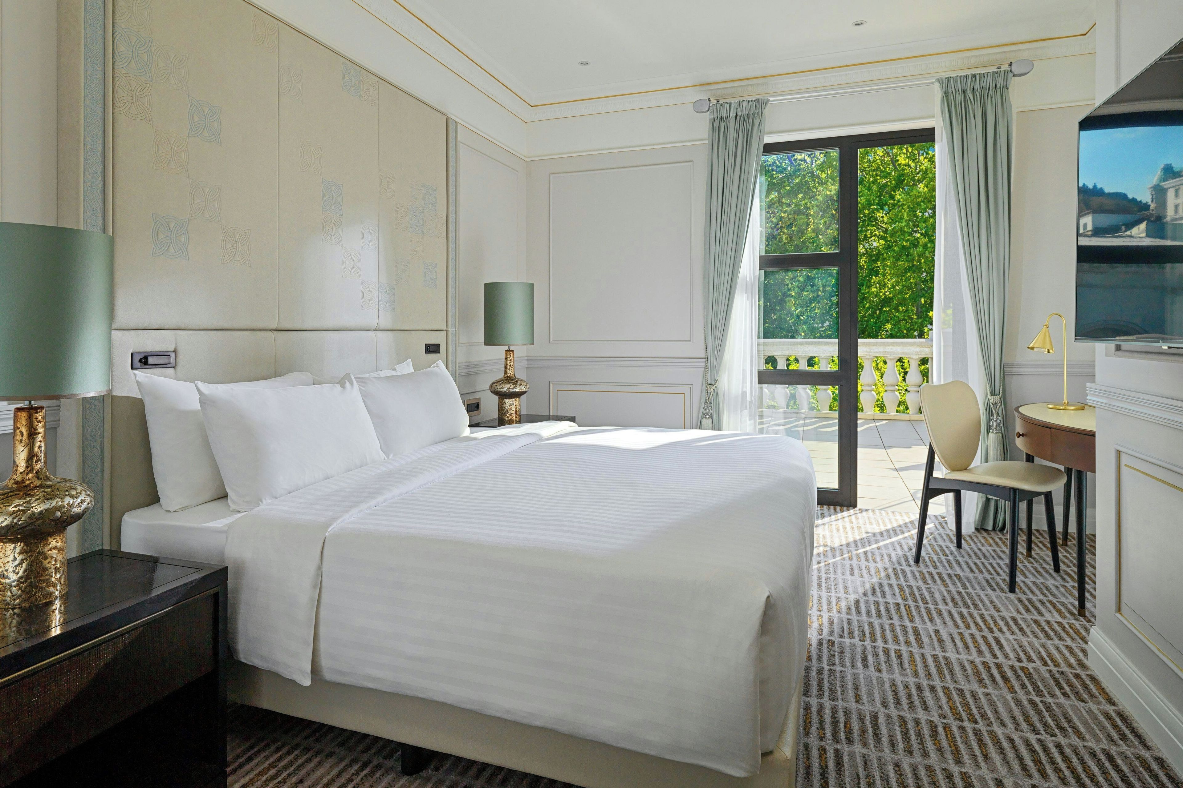 A hotel room with a large bed, a desk, a chair, and a view of greenery through the window.