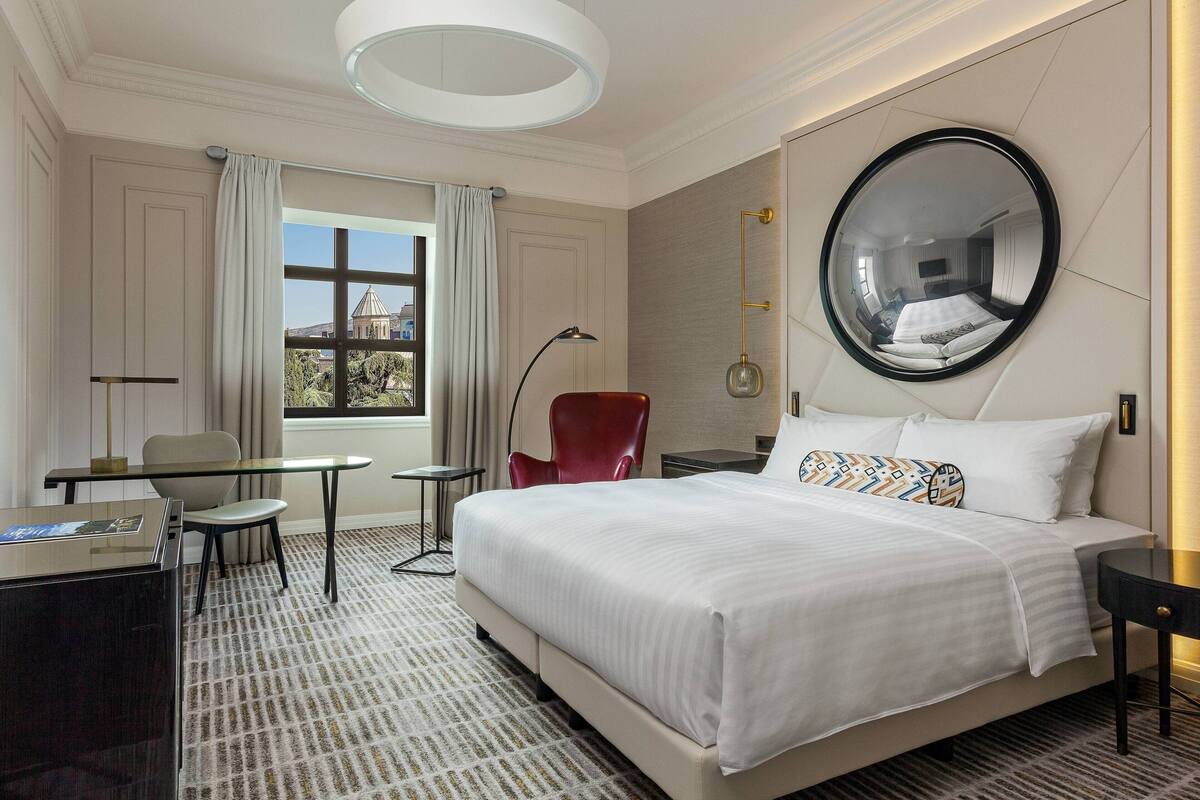 A hotel room with a large bed, a desk, a chair, a round mirror, and a window with curtains.