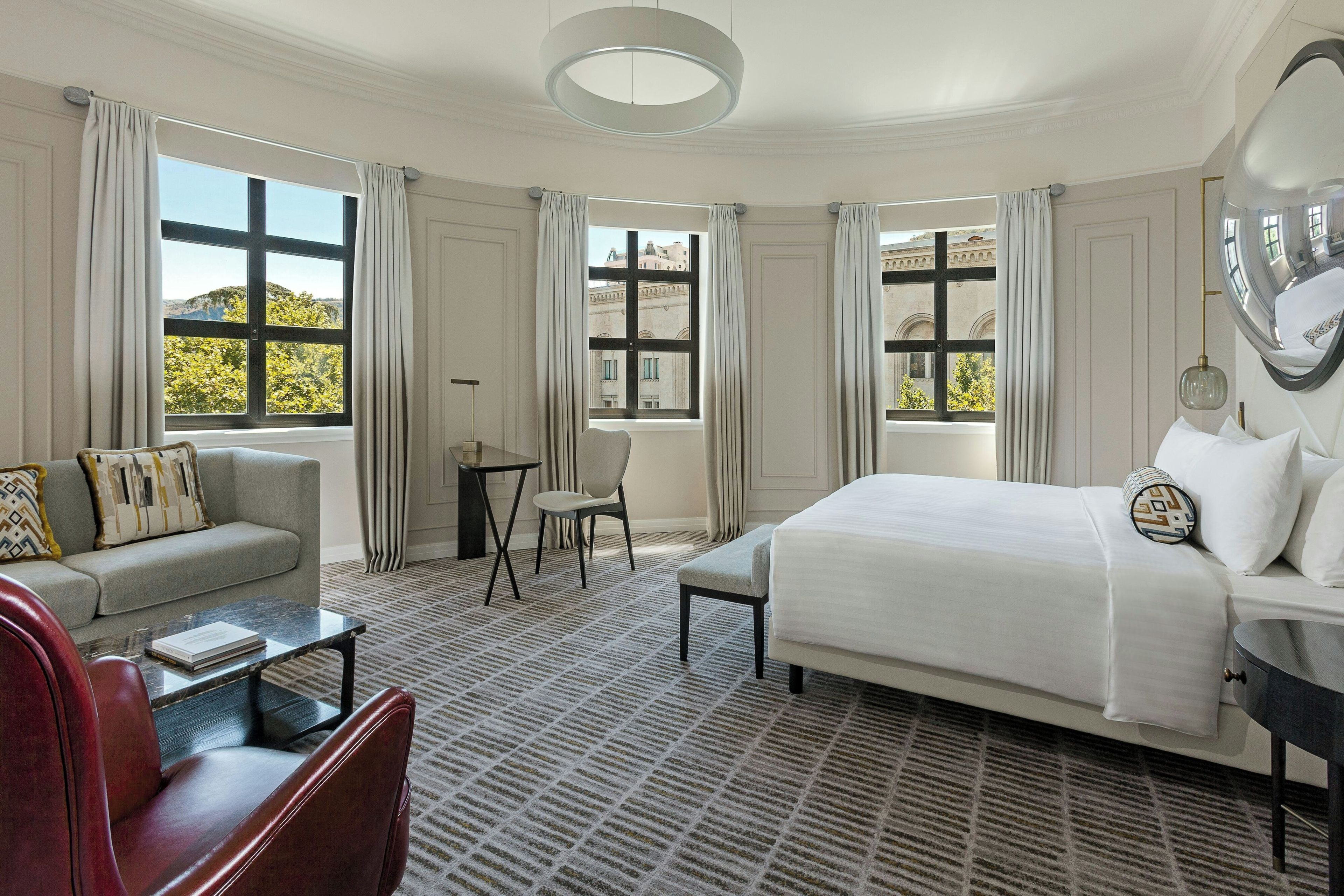 Habitación de hotel con cama, sofá, sillón, mesita y ventanas amplias con cortinas.