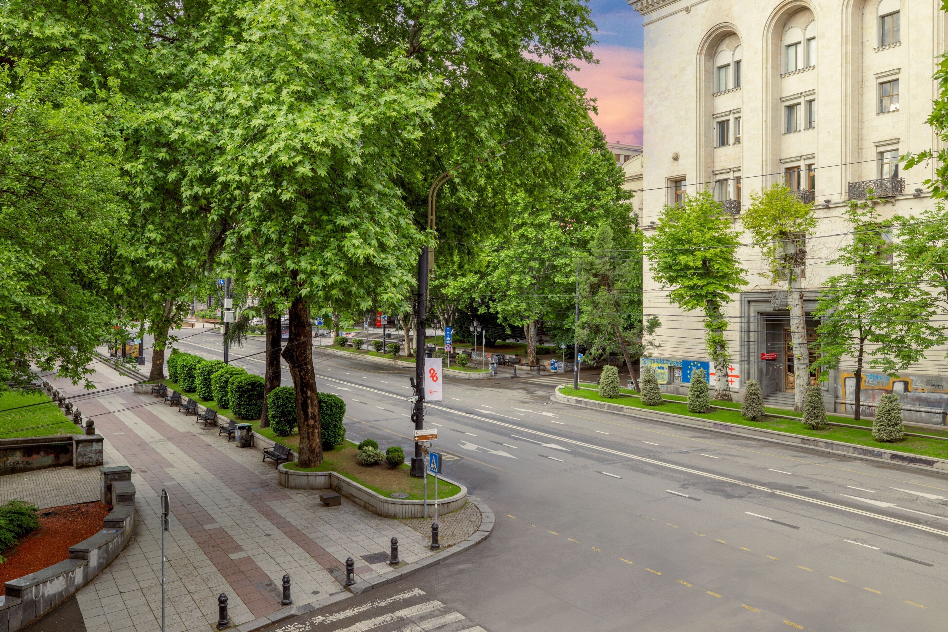 Une rue urbaine bordée d’arbres, avec un trottoir pour les piétons et un bâtiment doté de fenêtres en arc de cercle.