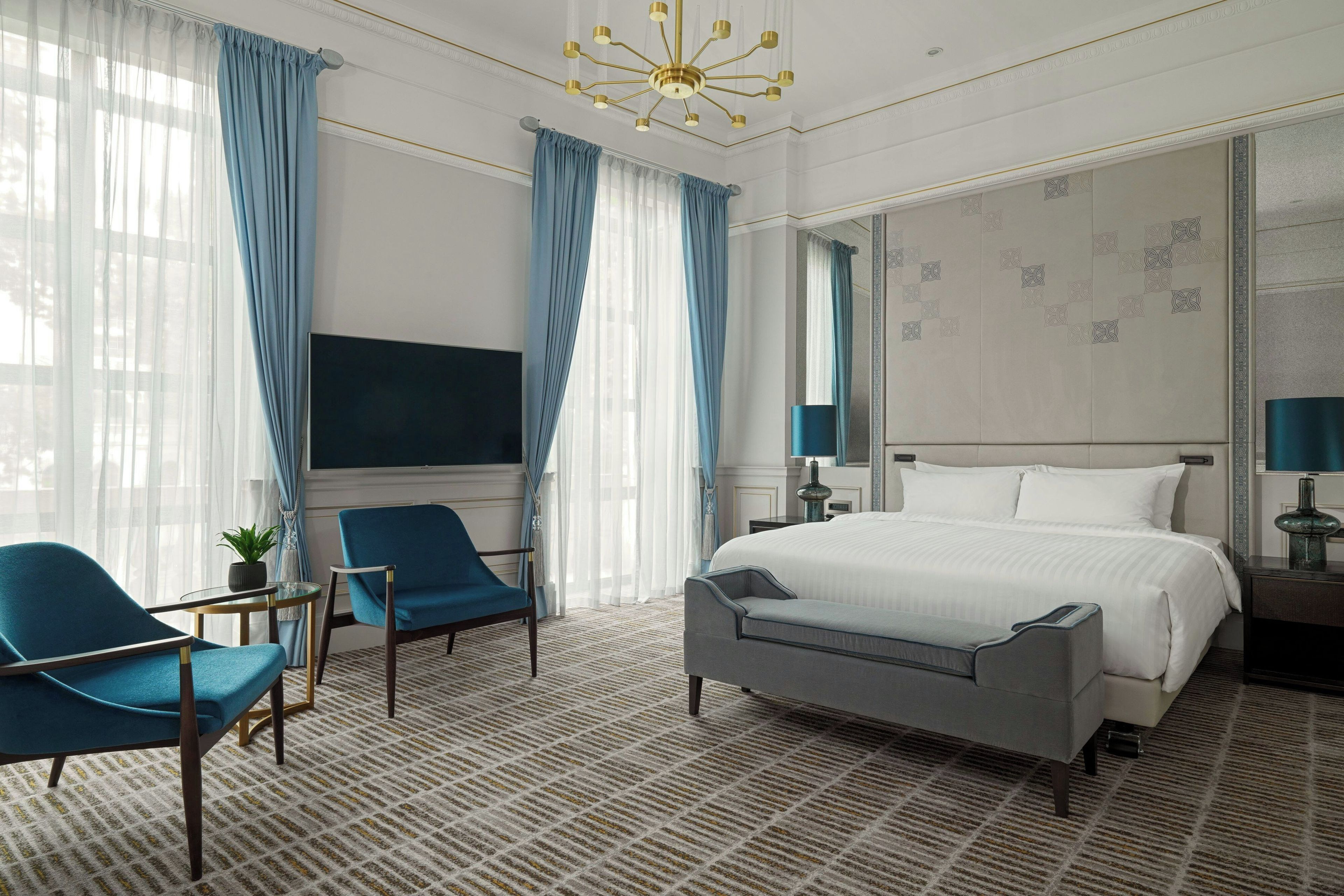 Quarto de hotel com uma cama grande, duas cadeiras, uma televisão e um candelabro.
