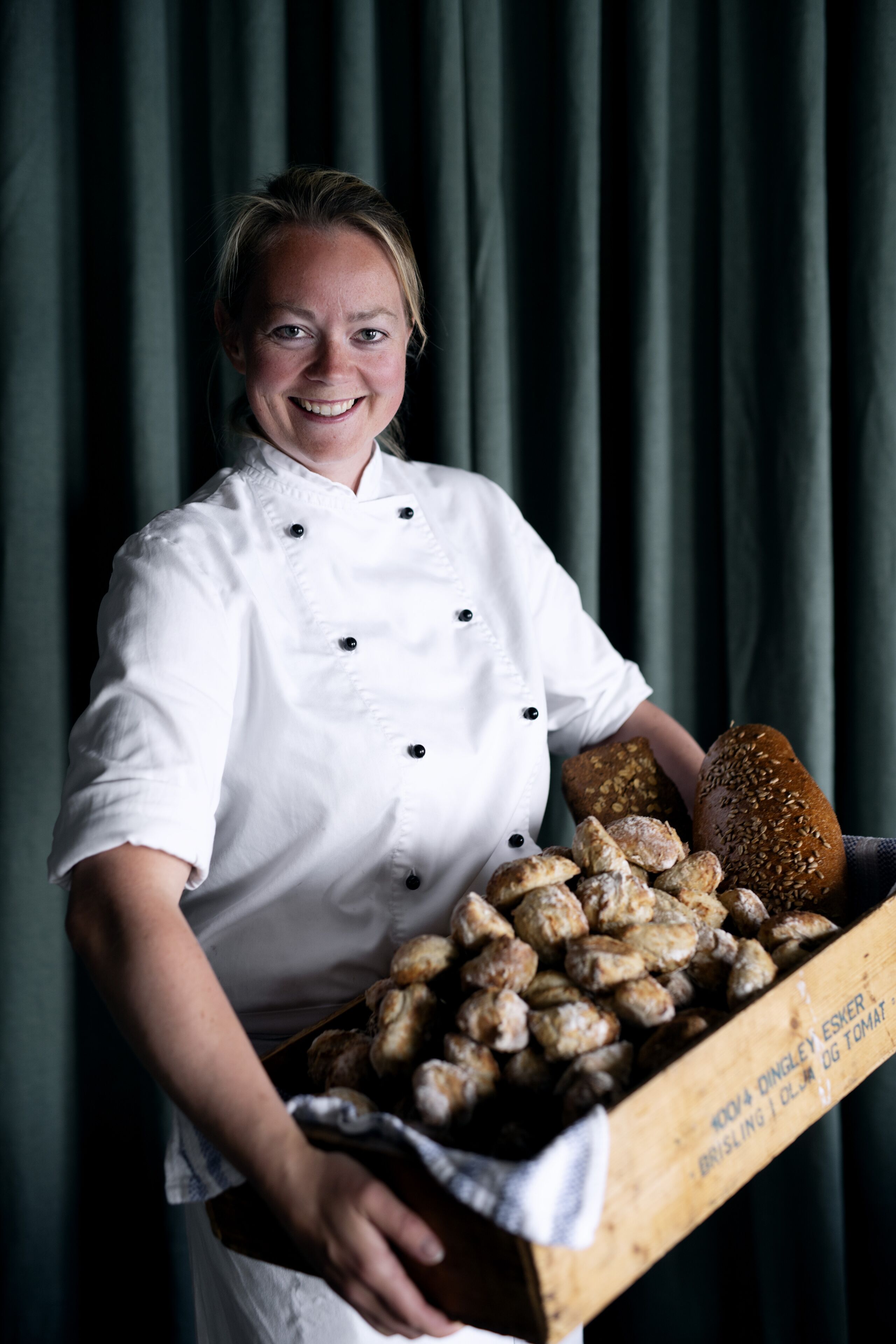 A chef in a white uniform holding a box of various breads and pastries.