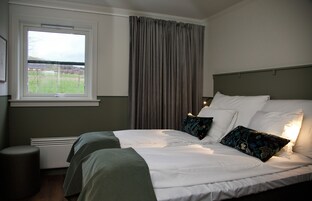Una habitación de hotel con una cama grande, una ventana con cortinas, un radiador y un taburete pequeño.