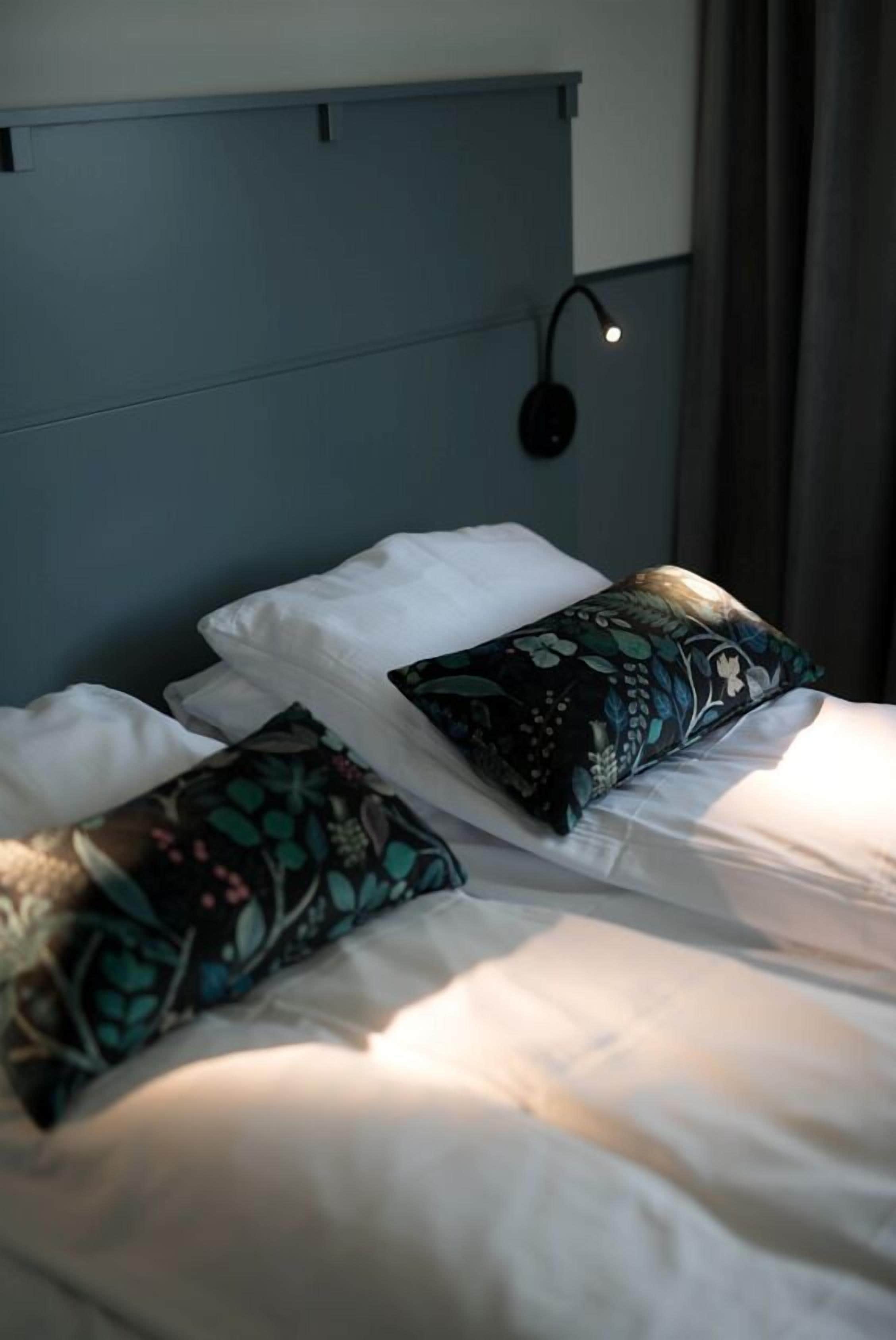 A neatly made bed with white linens and two decorative pillows, a wall-mounted light fixture, and a dark blue headboard.