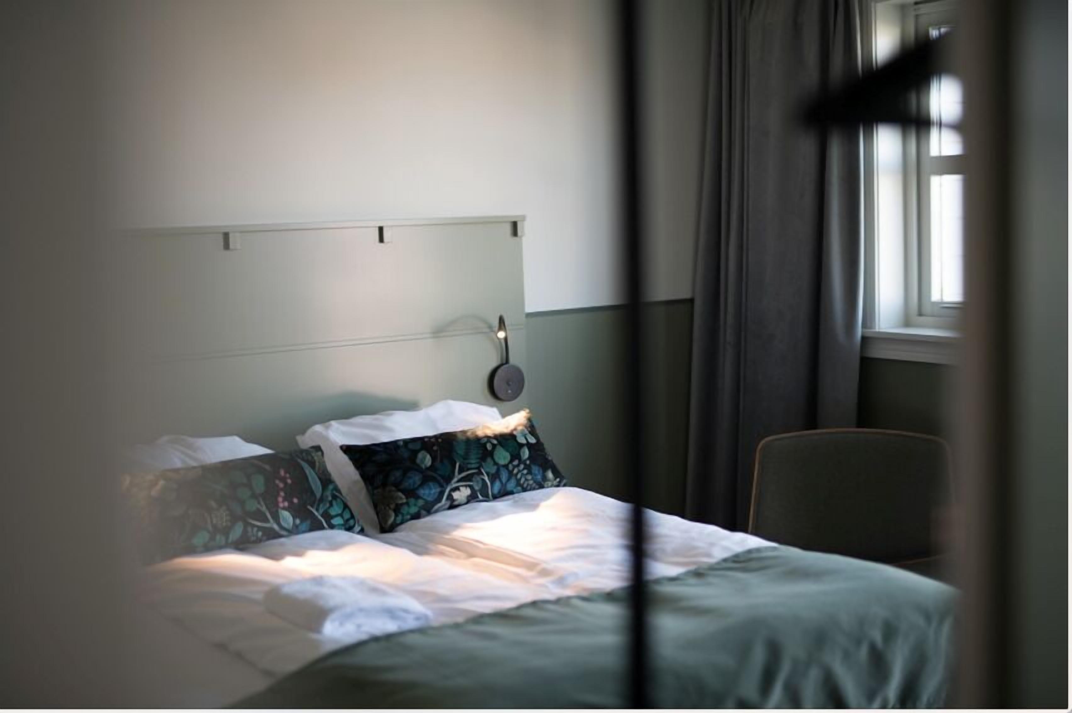 Una cama bien hecha con sábanas blancas y almohadas estampadas, una mesita de noche con lámpara, y una ventana con cortinas.
