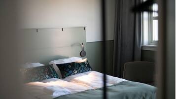 Una cama bien hecha con sábanas blancas y almohadas estampadas, una mesita de noche con lámpara, y una ventana con cortinas.