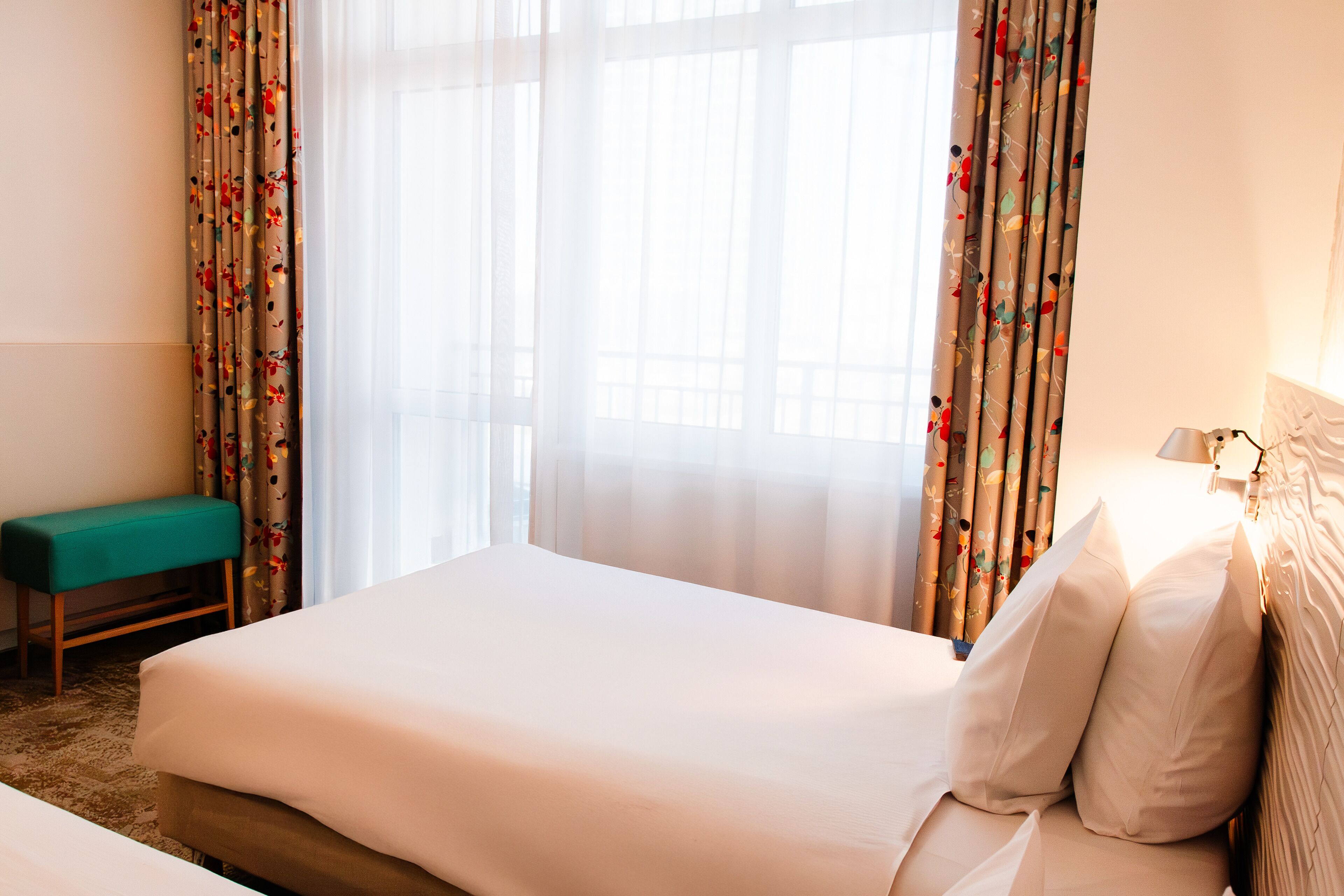 Habitación de hotel con una cama grande, un banco y cortinas translúcidas estampadas con motivos florales.