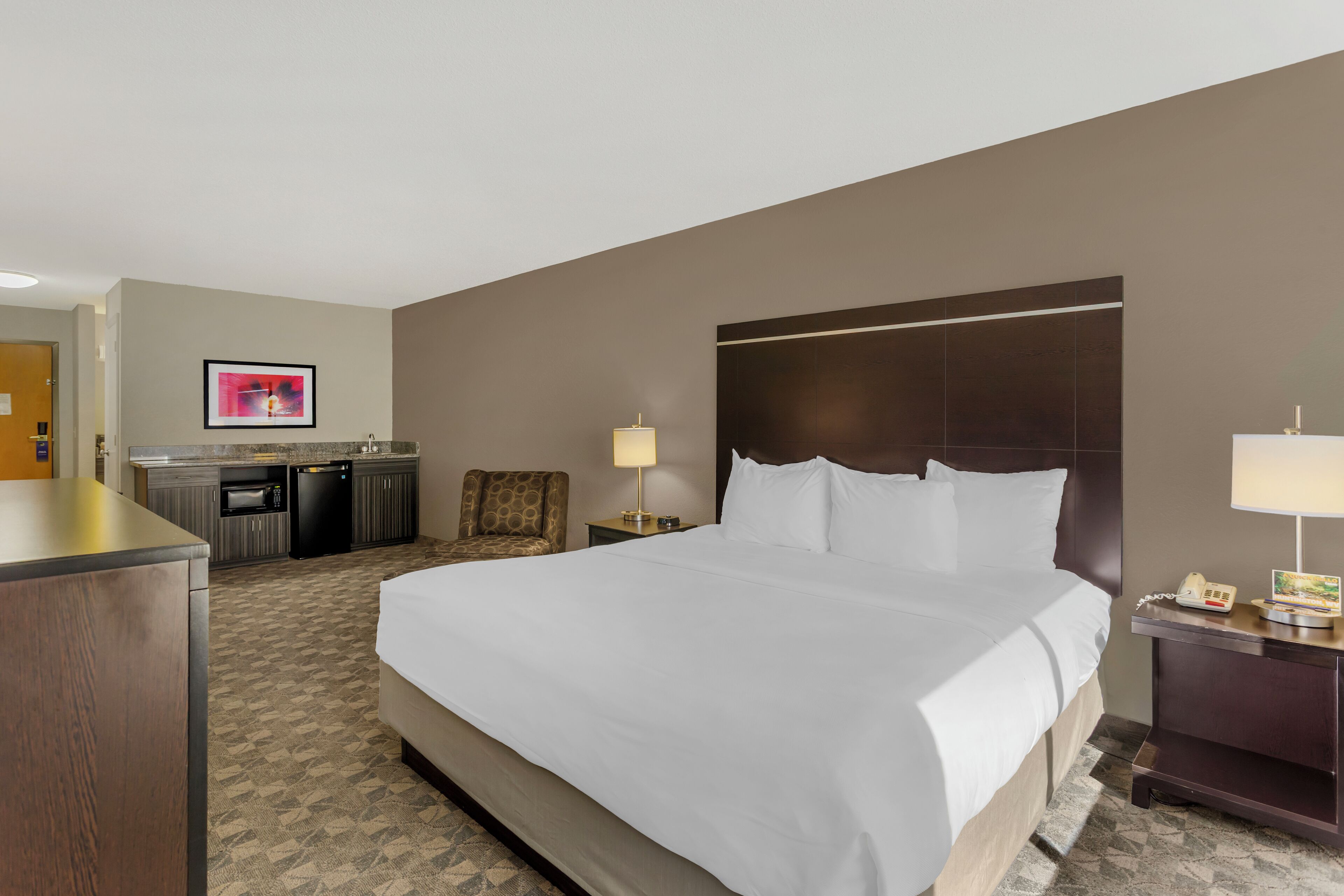 Habitación de hotel con una cama grande, un escritorio, una silla, una pequeña kitchenette y una mesita de noche con lámpara y teléfono.
