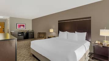 Habitación de hotel con una cama grande, un escritorio, una silla, una pequeña kitchenette y una mesita de noche con lámpara y teléfono.