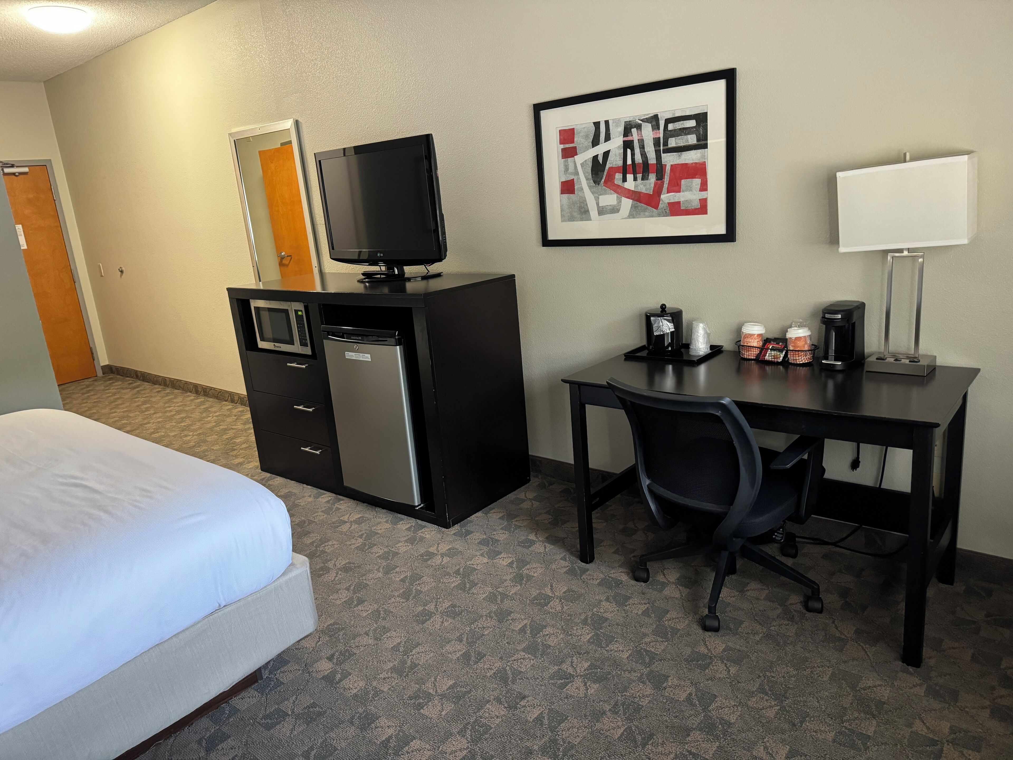 A hotel room with a bed, a desk with a chair, a microwave, a refrigerator, a television, a coffee maker, and a framed picture on the wall.