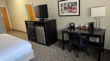 A hotel room with a bed, a desk with a chair, a microwave, a refrigerator, a television, a coffee maker, and a framed picture on the wall.