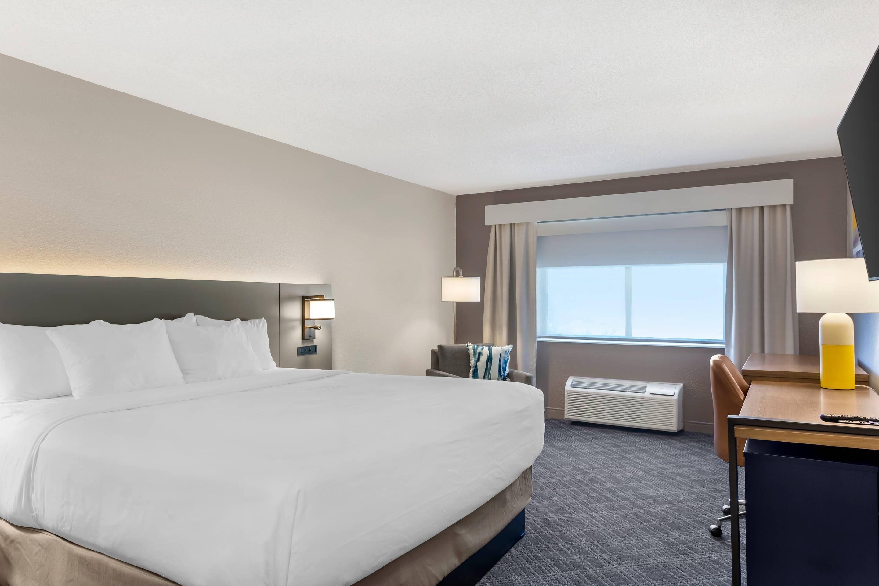 Una habitación de hotel con una cama grande, un escritorio con silla, una ventana con cortinas y un aire acondicionado instalado en la pared.