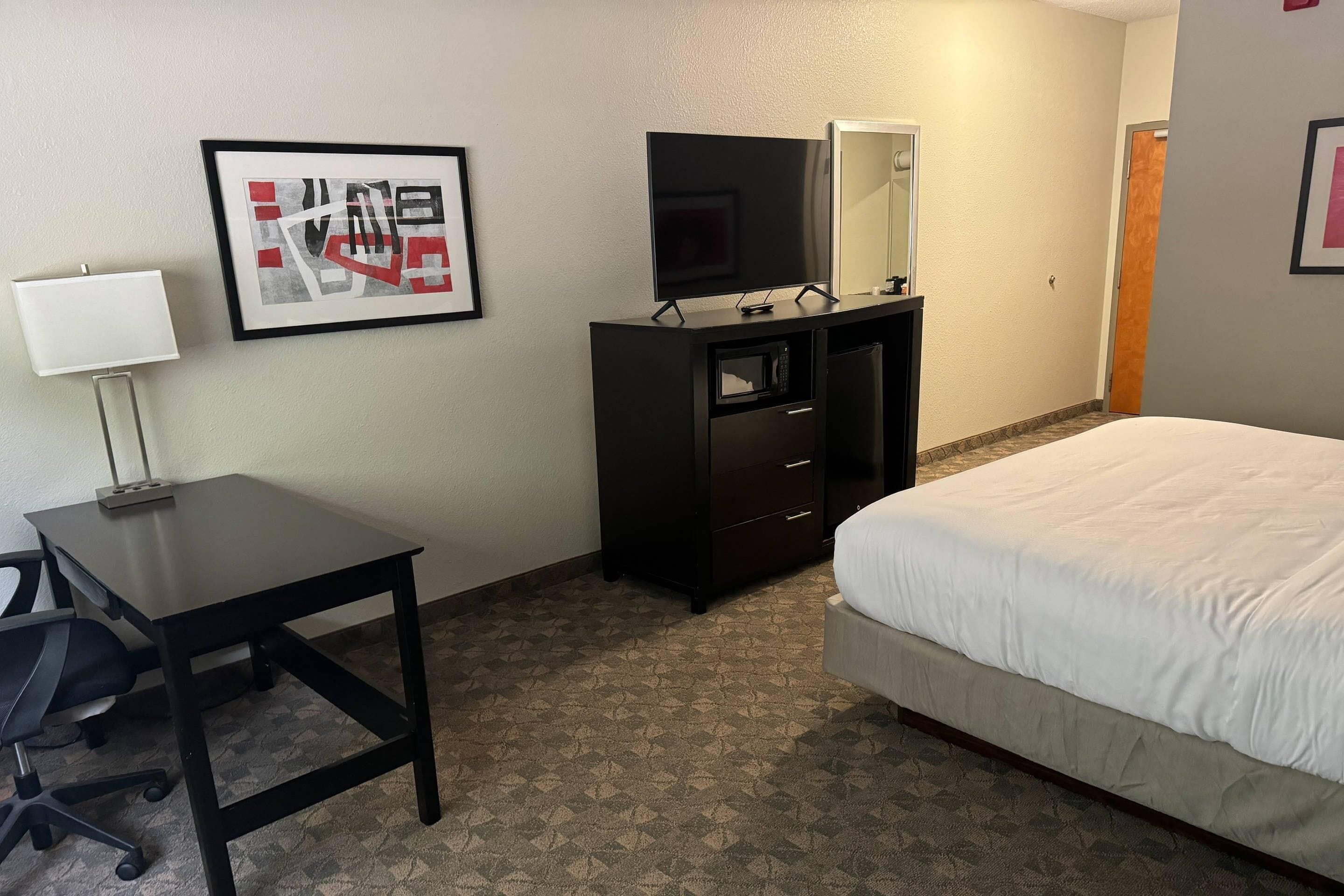 Una habitación de hotel con cama, escritorio con silla, cómoda, televisor de pantalla plana y un cuadro en la pared.
