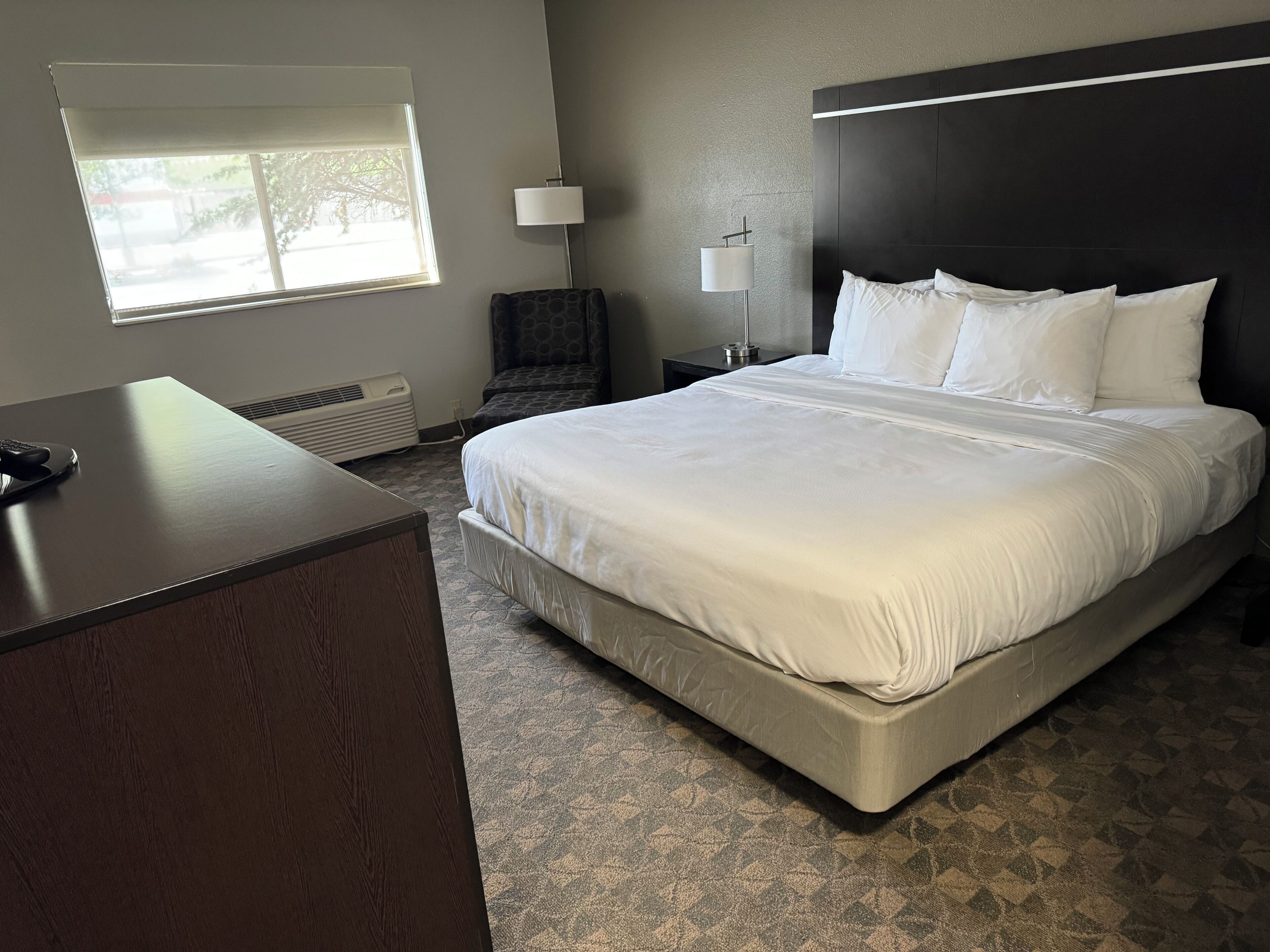 Ein Hotelzimmer mit einem großen Bett, einem Schreibtisch, einem Sessel, einem Nachttisch mit Lampe und einem Fenster mit Jalousien.