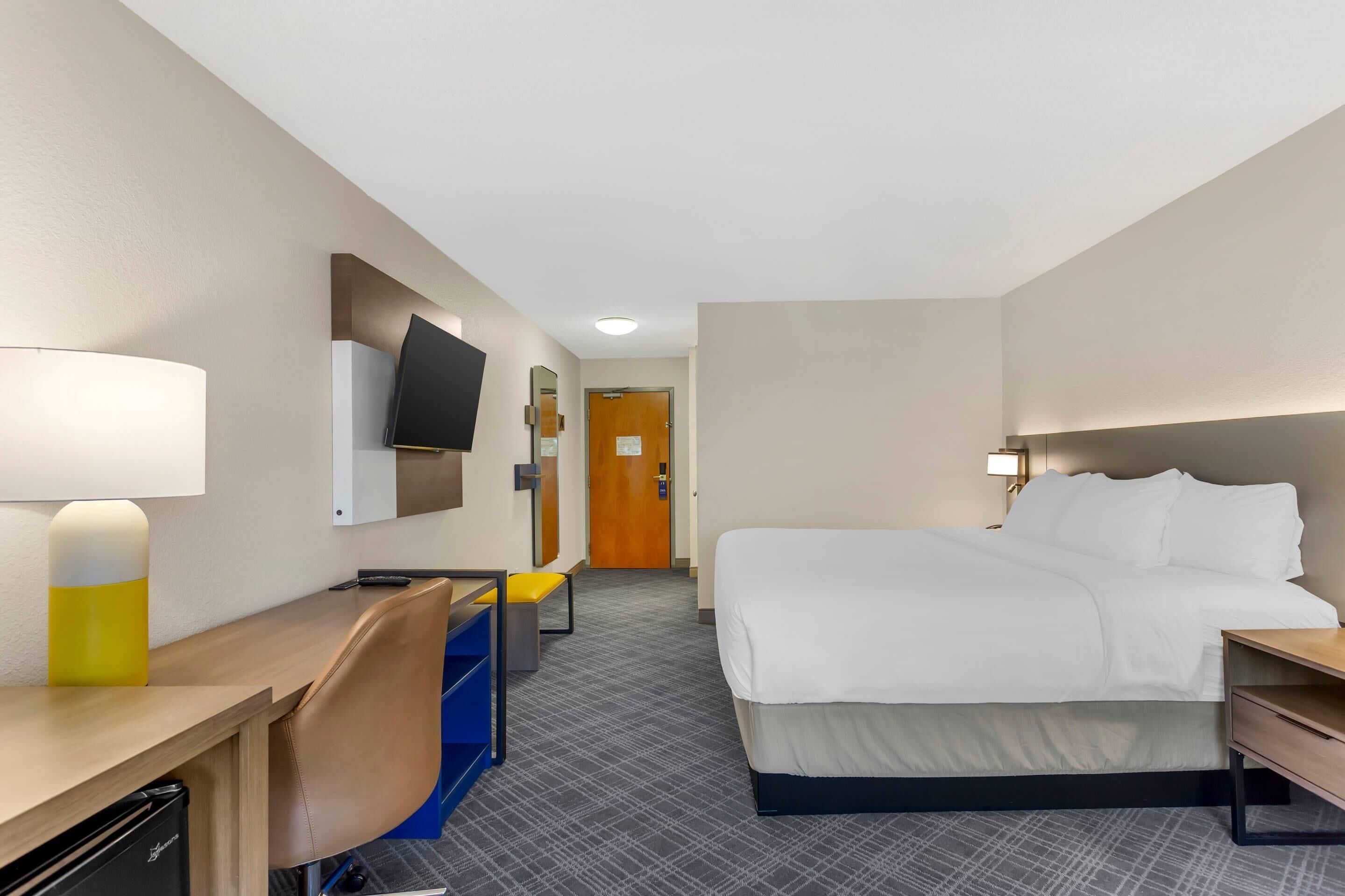 Ein Hotelzimmer mit Bett, Schreibtisch, Stuhl, Fernseher und einer Tür im Hintergrund.