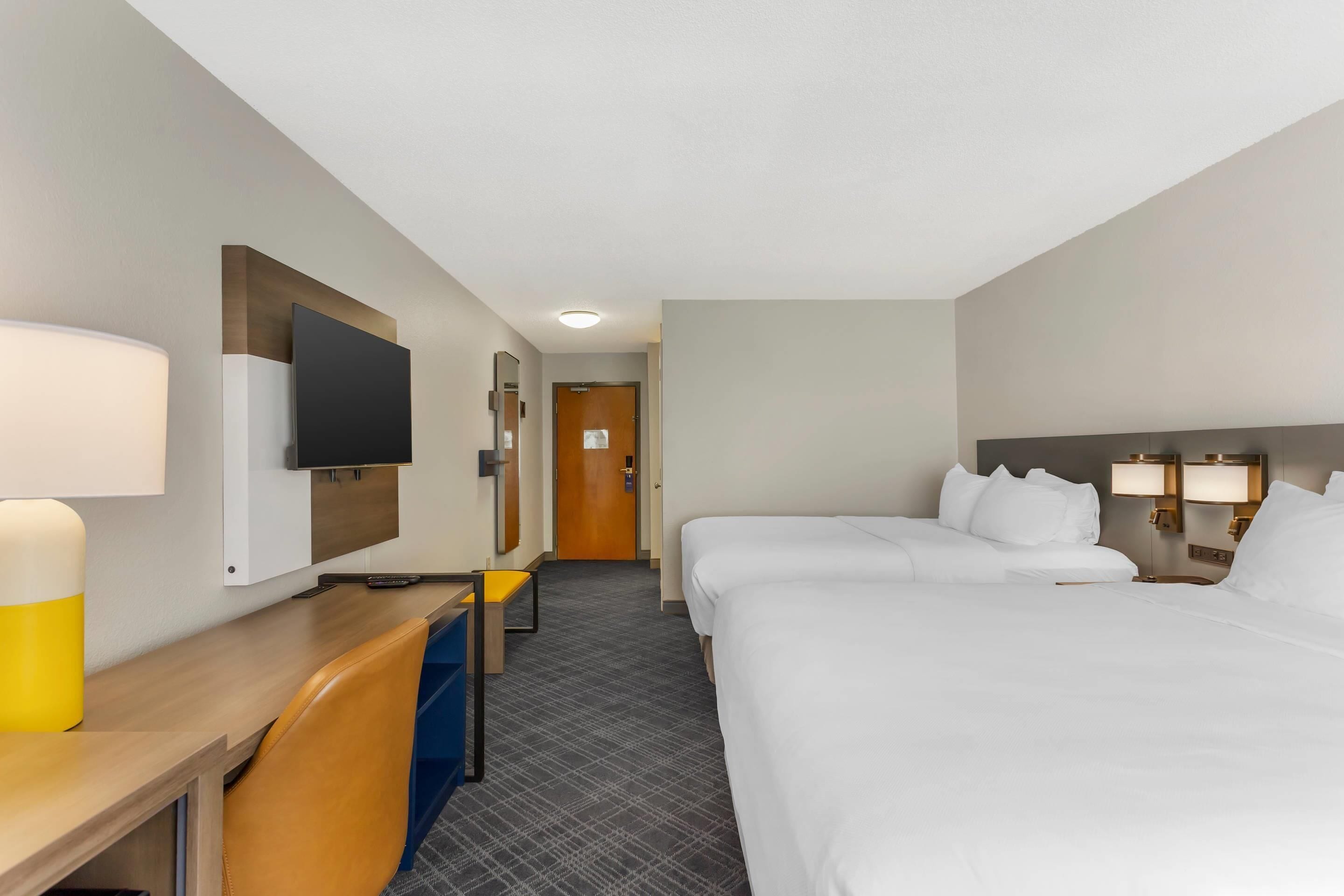 A hotel room with two beds, a desk with a chair, a TV, and a door leading to another room.