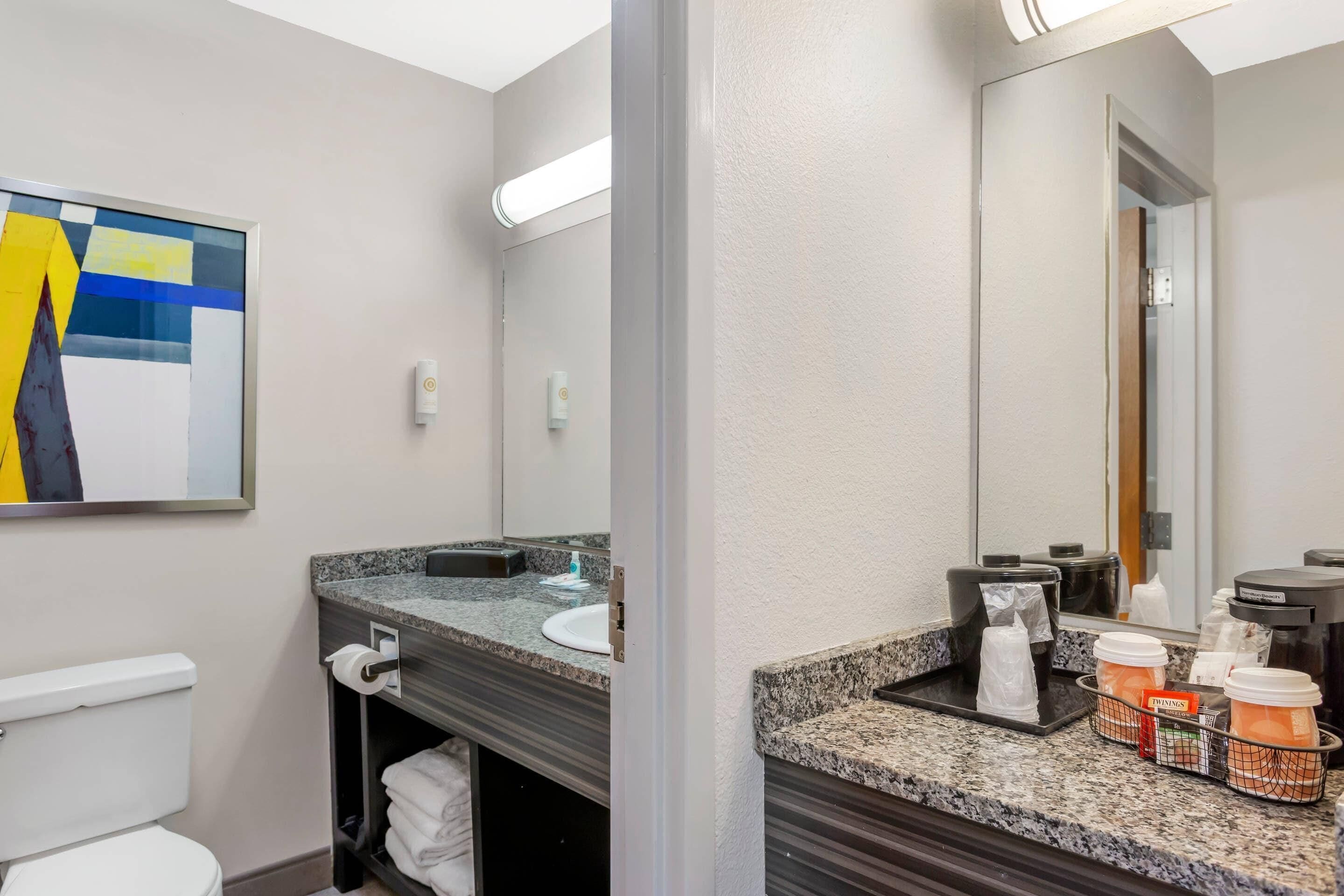 A modern bathroom with a granite countertop, a toilet, a sink, and a mirror. There are toiletries and a coffee maker on the counter.