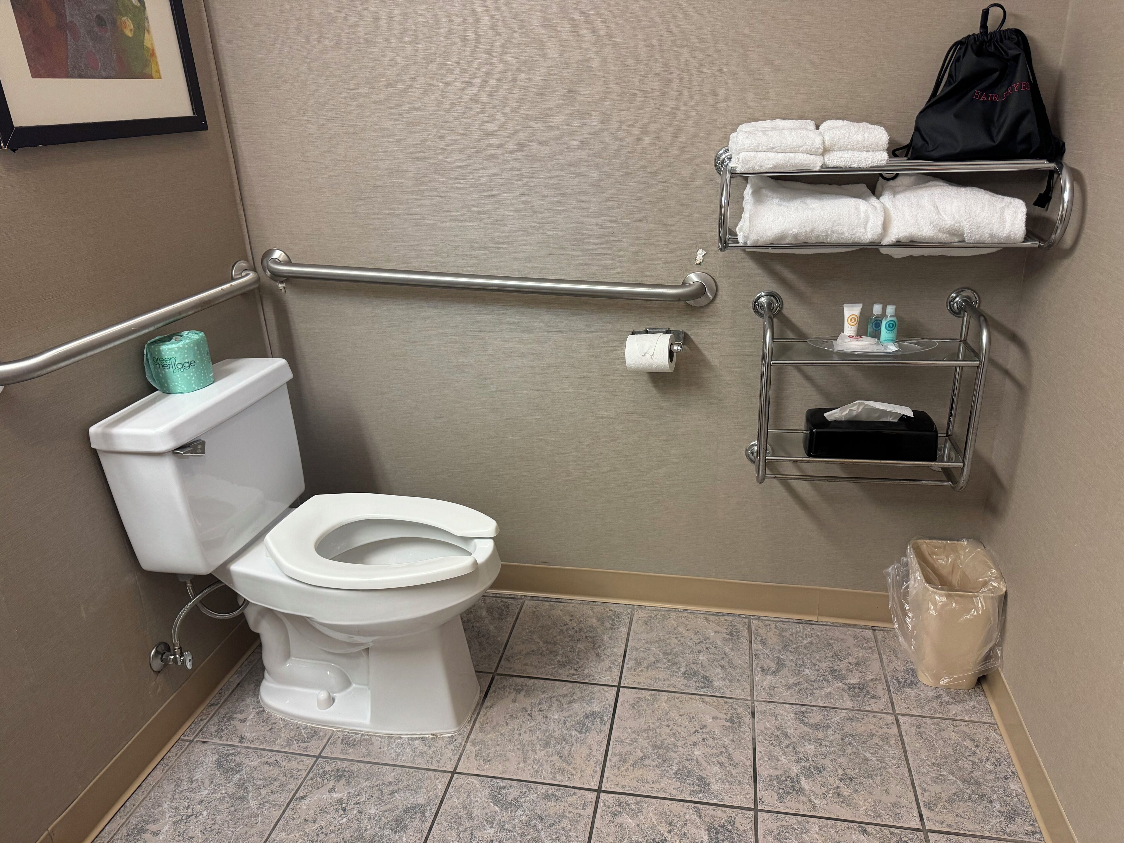 Une salle de bain équipée de toilettes, d’une barre d’appui, d’une étagère avec des serviettes et d’un sac contenant des articles de toilette.