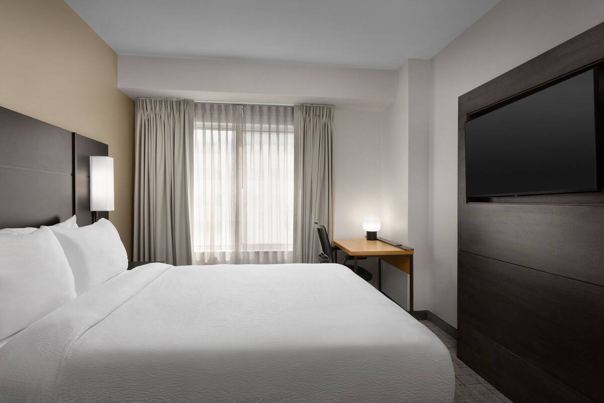 A hotel room with a large bed, a desk, a flat-screen TV, and a window with curtains.