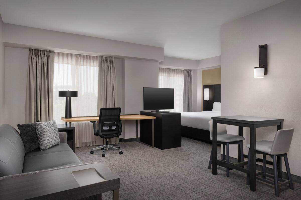 A modern hotel room with a sofa, desk, TV, and a small dining area.