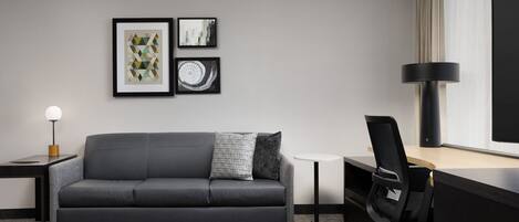 Een moderne hotelkamer met een grijze bank, een klein bijzettafeltje met een lamp, een bureau met een stoel, en kunst aan de muur.