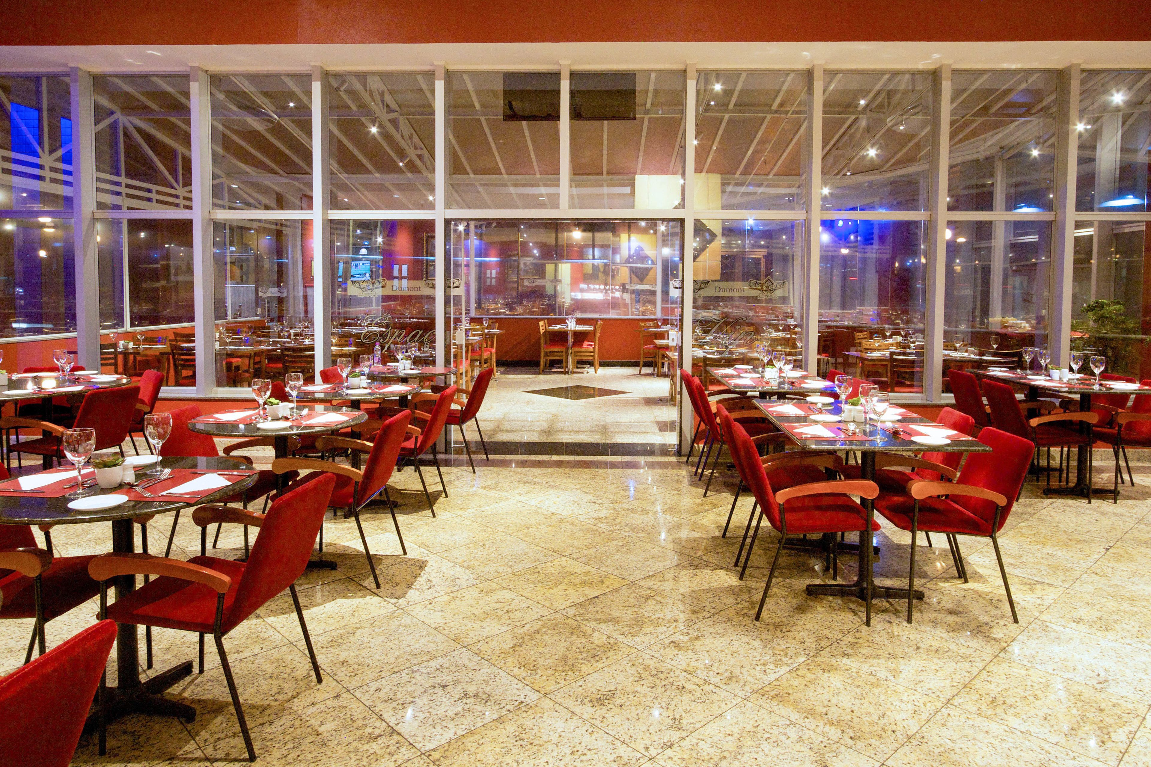 Un restaurante espacioso con techos altos, ventanas amplias y sillas rojas. Las mesas están ordenadas con copas de vino y menús.