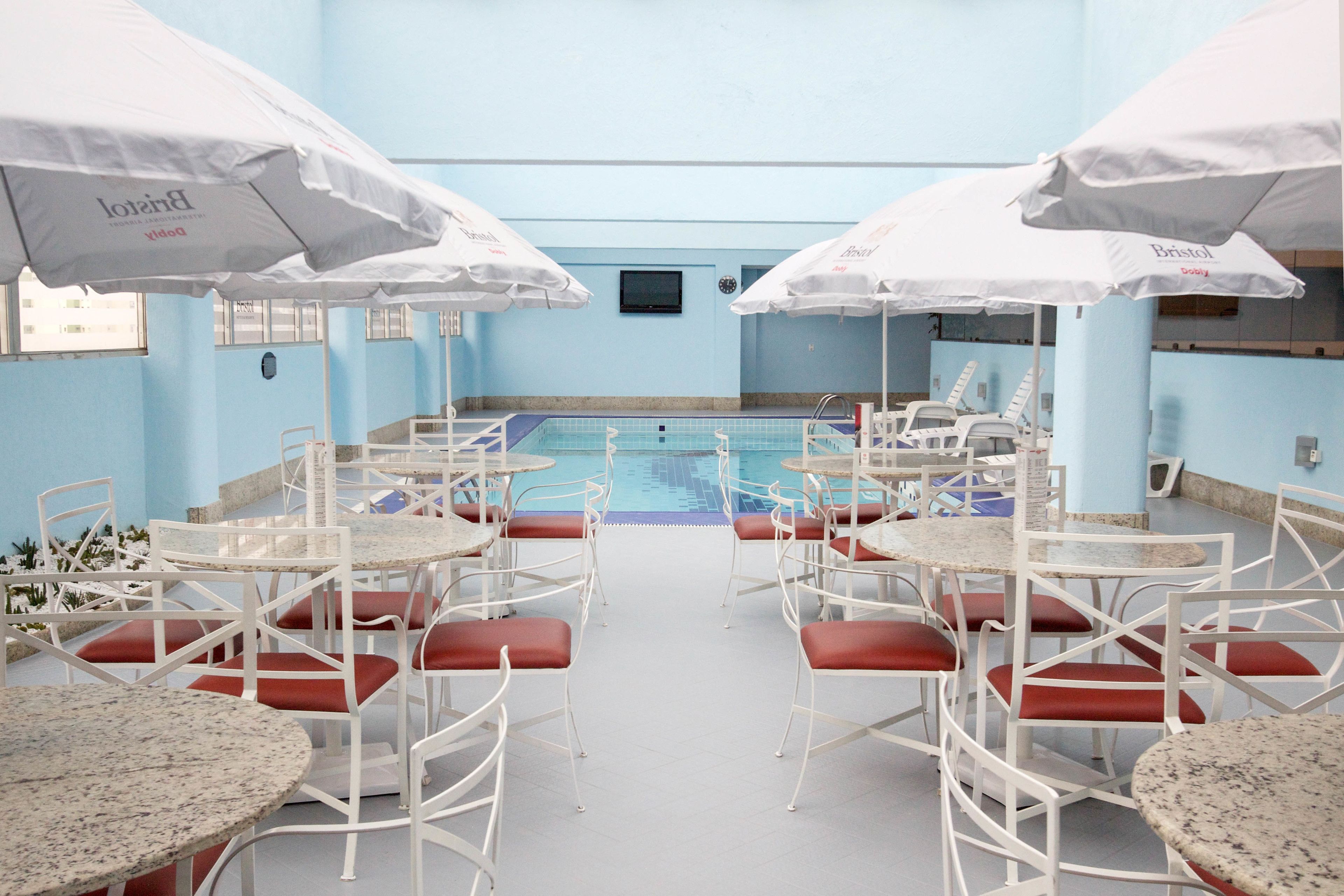 Un área de piscina con sillas y mesas blancas, sombrillas y una piscina con baldosas.