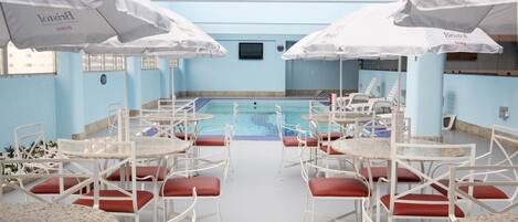 Un área de piscina con sillas y mesas blancas, sombrillas y una piscina con baldosas.