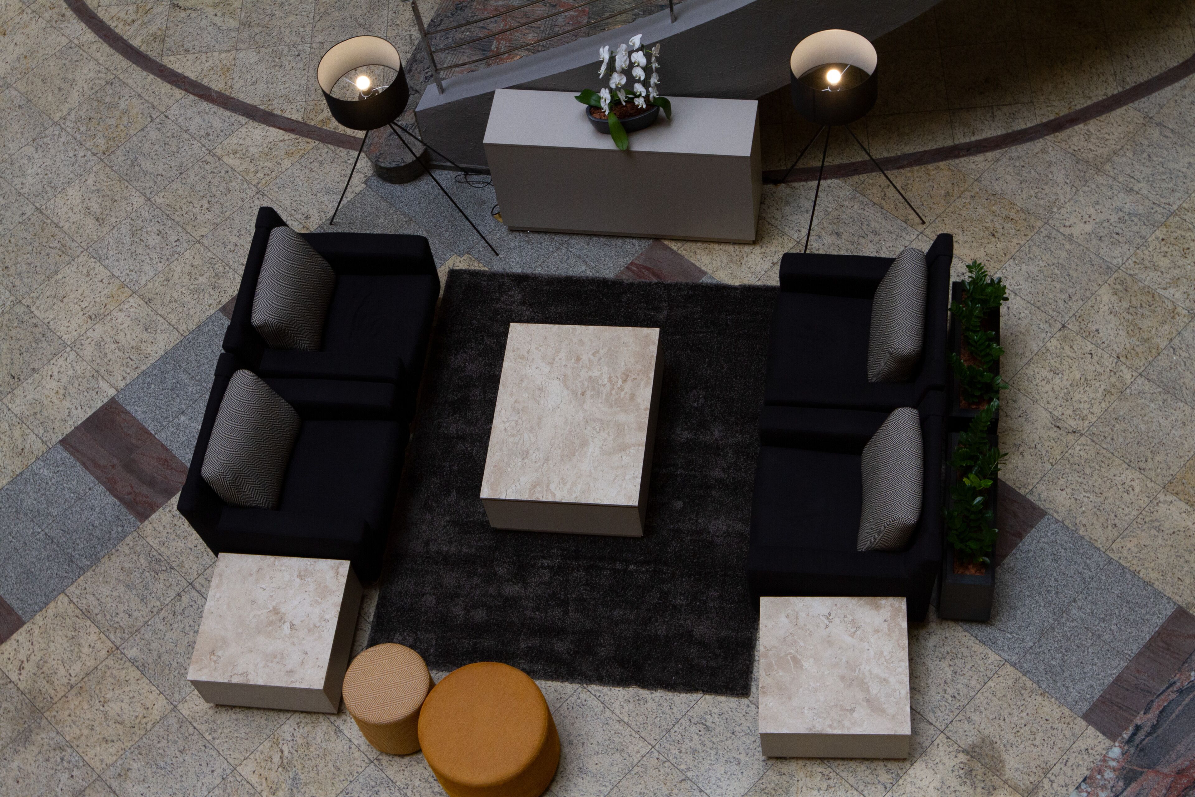 A lobby with a black sofa set, two side tables, a central coffee table, and a reception desk with a potted plant.